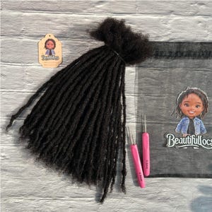 Puede incluir: Un conjunto de rastas de color marrón oscuro, atadas juntas, con dos ganchillos rosas. También hay una pequeña etiqueta y una pegatina con una ilustración de dibujos animados y el texto "Beautifullocs". Los artículos están dispuestos sobre una superficie de madera gris y blanca.