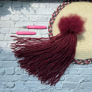 May include: A collection of burgundy dreadlocks with a matching fluffy top, displayed on a beige and blue braided mat. Two pink crochet hooks, each labeled "0.5mm", are positioned above the dreadlocks against a white brick background.