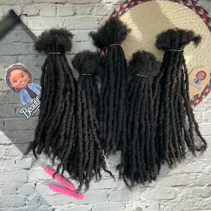 Pode incluir: Seis pacotes de dreadlocks de cabelo humano preto, cada um com uma ponta em laço. As dreadlocks estão dispostas em forma de leque, com as pontas das dreadlocks apontando para a parte inferior da imagem. As dreadlocks estão sobre uma superfície branca. A imagem também inclui dois ganchos de crochê cor-de-rosa.