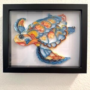 Op de afbeelding: Een ingelijst papieren kunstwerk van een kleurrijke zeeschildpad met blauwe, oranje en gele papieren stroken. De schildpad zwemt op een witte achtergrond.
