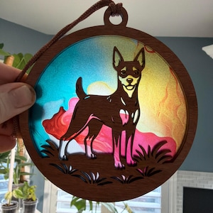 May include: A round, wooden dog ornament with a brown frame and a colorful background. The dog is brown with white markings, and the background features shades of blue, red, and yellow. A brown string is attached for hanging.