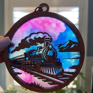 Op de afbeelding: Een ronde houten ornament met een treinontwerp. De trein staat tegen een kleurrijke achtergrond van roze, blauw en wit, met een bergketen in de verte. Het ornament heeft een bruine rand en een lus om op te hangen.
