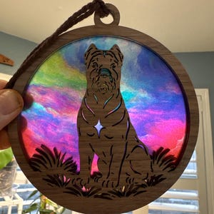 May include: A circular wooden dog ornament with a colourful, iridescent background. The dog is a silhouette, and the ornament has a brown cord for hanging. The background has shades of pink, purple, blue, and green.