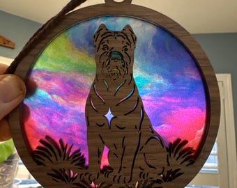 May include: A wooden dog ornament with a colorful, iridescent background. The dog is a silhouette, and the ornament has a brown cord for hanging. The background has shades of pink, purple, blue, and green.
