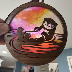 Pode incluir: Enfeite circular de madeira com um desenho de lontra cortado a laser flutuando na água. A lontra é marrom e a água é vermelho-alaranjada. O fundo é uma mistura de rosa, laranja e amarelo. Uma corda marrom está presa para pendurar. Mede cerca de 10 cm de diâmetro.