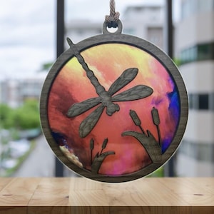 May include: A wooden ornament featuring a dragonfly silhouette against a colorful watercolor background. The background is a mix of pink, orange, yellow, and blue.