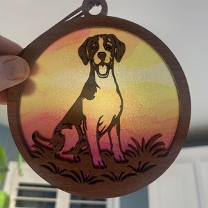 Pode incluir: Enfeite redondo de madeira com um cão marrom e branco contra um fundo de pôr do sol colorido. O cão está sentado em frente a uma área gramada. O enfeite tem uma moldura de madeira marrom escura e uma corda para pendurar.
