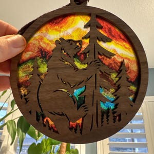 May include: A circular wooden ornament with a bear silhouette hugging a tree, set against a vibrant sunset. The brown ornament has a hanging cord. The design includes trees and a bear, creating a nature-themed decoration.