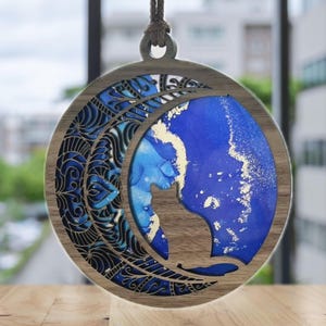 Könnte beinhalten: Ein Holz-Anhänger in Form eines Halbmondes mit einem blauen und goldenen Harz-Einlegearbeiten. Der Mond ist mit einem schwarzen und blauen Mandala-Design verziert. Der Anhänger hängt an einer braunen Schnur.