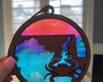 May include: A circular wooden ornament with a crab design and a layered, colourful background. The ornament has a dark brown wooden frame with a crab silhouette and a state outline. The background has shades of blue, purple, and orange.