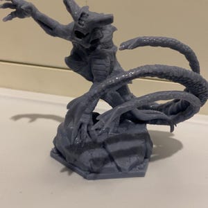 Puede incluir: Una miniatura impresa en 3D gris de una criatura con una cola larga y garras. La criatura está de pie sobre una base rocosa.
