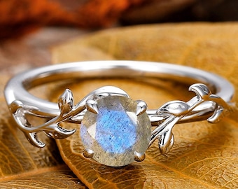 Op de afbeelding: Een zilveren ring met een ronde, iriserende blauwe edelsteen. De ring heeft een delicaat wijnstokontwerp op de band, met bladeren aan weerszijden van de edelsteen. De ring ligt op een bruin blad.
