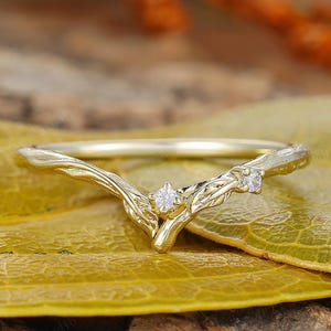 May include: A delicate gold ring with a V-shaped design. The ring features a branch-like motif with two small, clear gemstones. The ring is set against a yellow leaf and a wooden surface.