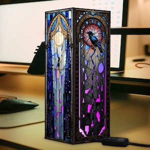 Puede incluir: Lámpara decorativa de vidrieras con diseño gótico. La lámpara presenta una combinación de colores azul y morado con un diseño de cuervo y luna. La lámpara es rectangular y tiene un cable de alimentación.