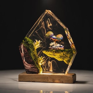 May include: A clear, geometric resin sculpture containing a miniature landscape with a mossy rock formation, small white plants, and a colorful tree on a floating mossy island, all mounted on a rectangular wooden base with internal lighting.