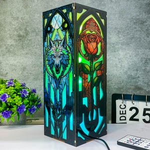 Può includere: Lampada decorativa rettangolare con pannelli in stile vetrata. Un pannello presenta un lupo blu e bianco, l'altro una rosa rossa. La lampada ha una cornice nera ed emette una luce verde e blu. È visibile un telecomando.