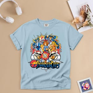 May include: Pale blue t-shirt with a vibrant ThunderCats graphic. The design showcases the ThunderCats logo and various characters in dynamic poses. The shirt is made of a soft fabric and has short sleeves.