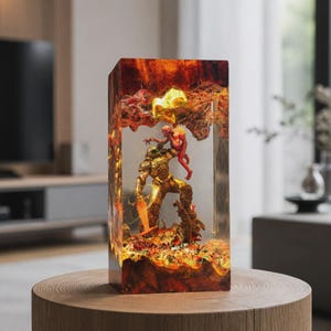 May include: A rectangular resin lamp featuring a detailed Doom Slayer figure in gold armour lifting a red demon. The base and top are encased in a fiery orange and red textured resin block, creating a glowing, molten effect around the central scene.