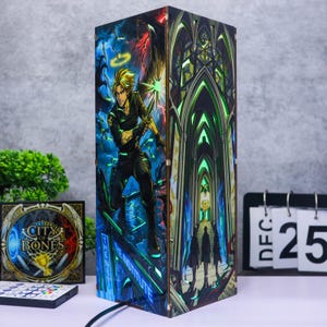 以下が含まれることがあります： City of Bonesの鮮やかなイラストが描かれた長方形の木製ランプ。片面には光る天使の輪をつけた金髪のキャラクターが描かれ、もう片面には緑色の光のアクセントが施されたゴシック様式の建築的なインテリアが描かれています。