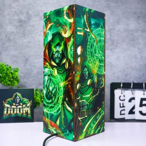 May include: A rectangular wooden lamp featuring a vibrant graphic print of the comic book character Dr. Doom in a green hooded cloak, casting glowing green magical energy circles from his hands. The lamp emits light through laser-cut details.