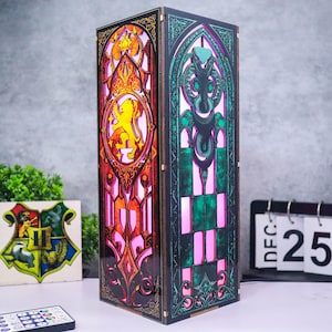 Può includere: Lampada decorativa rettangolare in stile vetrata. Presenta disegni intricati con motivi di leone e serpente, illuminati con luce rosa e arancione. La lampada in legno è dotata di telecomando. Misure in centimetri.