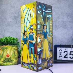 May include: A rectangular wooden lamp featuring multi-layered cutouts of Snow White and the Prince. The front panel shows the characters in a forest, while the side panel depicts Snow White indoors. The lamp emits a soft blue light from within.