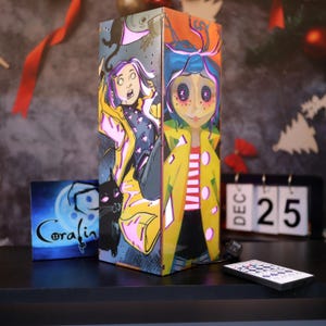 Pode incluir: Lâmpada retangular com ilustrações do filme "Coraline". A lâmpada apresenta personagens em amarelo e preto, um gato preto e a palavra "Coraline" em uma pequena pintura quadrada. Um controle remoto e um calendário mostrando 25 de dezembro também são visíveis.