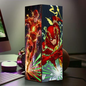 May include: A rectangular lamp featuring comic book art of the Flash in a running pose and another image of the Flash. The lamp has a red and yellow color scheme, with a cityscape background. The lamp is on a desk.
