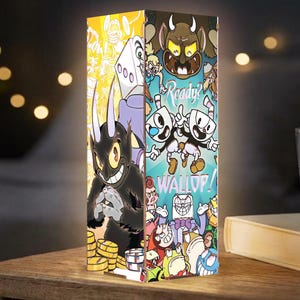 以下が含まれることがあります： 側面にカラフルなカップヘッドのゲームグラフィックが描かれた長方形のランプ。片面には金貨を持った黒い角のあるキャラクターが描かれ、もう片面には「Ready? WALLOP!」という文字と様々なゲームキャラクターが明るいシーンで描かれています。