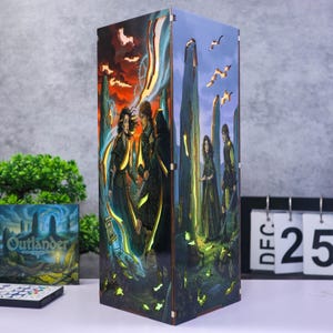 Puede incluir: Una lámpara rectangular de madera con ilustraciones coloridas de personajes en un paisaje con piedras erguidas. El panel frontal muestra una escena dramática con un hombre y una mujer, el panel lateral muestra figuras cerca de piedras.