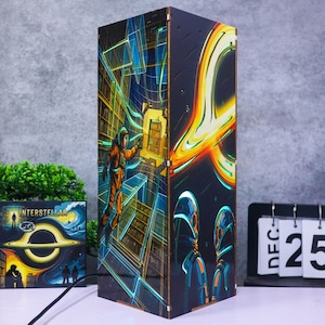 May include: A rectangular lamp featuring vibrant, multi-colored illustrations inspired by the film Interstellar. One side depicts an astronaut in an orange suit floating in a library, while the other side shows a glowing black hole and two figures.