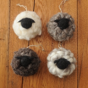 May include: Four felt sheep ornaments, each with a black felt face. Two sheep are white, one is grey, and one is brown.