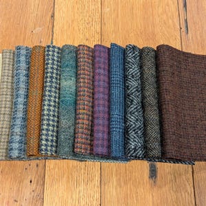 Op de afbeelding: Een verzameling van 14 stofstalen in verschillende kleuren en patronen, waaronder bruin, blauw, groen, oranje en beige. De stalen zijn in een rij op een houten oppervlak gerangschikt.