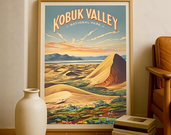 May include: A framed print of Kobuk Valley National Park. The artwork shows a desert landscape with sand dunes, mountains, and a sunset sky. The words "KOBUR VALLEY NATIONAL PARK" are printed at the top in an orange font. A white vase and stacked boxes are in the foreground.