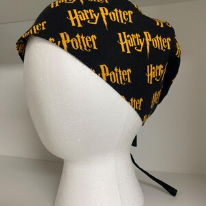 May include: Black scrub cap with the words "Harry Potter" in gold lettering. The cap is tied in the back with black ties. The cap is displayed on a white foam head.