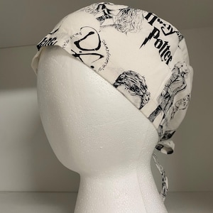 May include: White fabric cap featuring black illustrations and text from the Harry Potter series. The cap is tied at the back and is displayed on a white mannequin head. The text includes "Harry Potter" and "Dumbledore's Army".