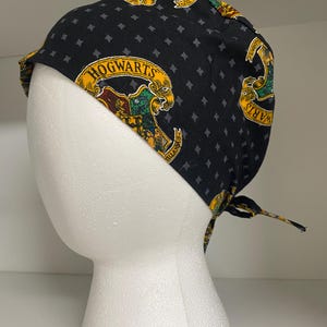 May include: Black surgical scrub cap with a repeating pattern of small gray diamonds and the Hogwarts crest in yellow, green, and maroon. The cap is tied in the back and is displayed on a white foam head.