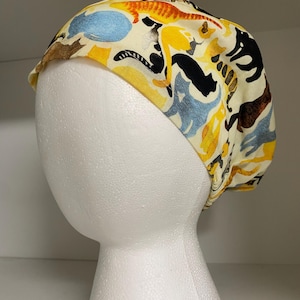 Può includere: Un cappello chirurgico fantasia con un motivo ripetuto di gatti colorati in giallo, nero, blu e marrone su sfondo crema. Il cappello è esposto su un manichino di schiuma bianca, che ne mostra la forma e il design.