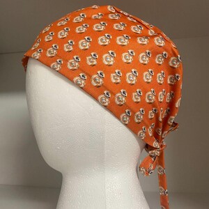 Może przedstawiać: Pomarańczowa chusta z tkaniny z powtarzającym się wzorem droidów BB-8. Nakrycie głowy jest zawiązane z tyłu i jest prezentowane na białej głowie manekina. Tkanina ma lekki połysk.