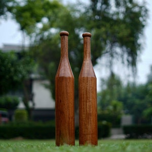 Puede incluir: Dos mazas indias de madera, de pie sobre una superficie cubierta de hierba. Las mazas son de color marrón oscuro, con una parte superior redondeada y un cuerpo largo y delgado. El fondo está borroso, con árboles y follaje verdes.