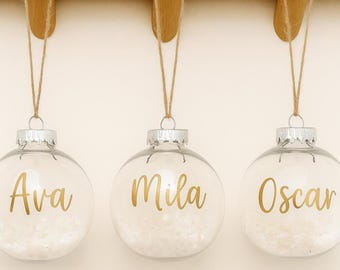 May include: Three clear glass Christmas ornaments with silver caps and gold lettering spelling out the names "Ava", "Mila", and "Oscar". Each ornament is filled with white decorative material and suspended by twine.