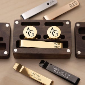 Może przedstawiać: Kolekcja spersonalizowanych akcesoriów męskich, w tym spinki do mankietów, krawaty i drewniane pudełka prezentacyjne. Spinki do mankietów mają inicjały "AG", a krawaty są grawerowane napisami.