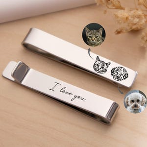 May include: Two silver-toned metal tie clips. One clip features a black engraved line drawing of a cat and a dog face. The other clip features the engraved cursive text I love you. Small circular photos of a cat and a dog are shown nearby.