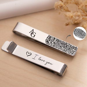 May include: Two silver-toned metal tie clips. One clip features the engraved letters AG and a detailed fingerprint design. The second clip displays a small heart symbol followed by the engraved cursive text I love you.