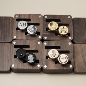 May include: A collection of personalized cufflinks displayed in individual wooden boxes. The cufflinks feature engraved initials, portraits, and names. The boxes are square with a dark wood finish and silver accents.