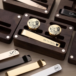 Puede incluir: Una colección de gemelos y pasadores de corbata personalizados en cajas de madera oscura. Los gemelos presentan una foto de una pareja, y los pasadores de corbata están grabados con fechas, nombres e iniciales. Algunos pasadores son dorados, negros y plateados.