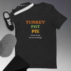 May include: Dark gray t-shirt with the words "TURKEY POT PIE" in orange and green, with "three of my favorite things" below. Black sneakers and a baseball cap are next to the shirt.