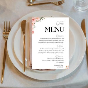 Puede incluir: Una tarjeta de menú blanca con detalles florales rosas, colocada sobre un plato blanco con cubiertos dorados. El menú presenta la palabra "MENU" en fuente grande y elegante, y enumera opciones de entrante, plato principal y postre. La fecha "07.07.20XX" también es visible.