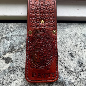 May include: A handcrafted, reddish-brown leather phone case with intricate floral and chain-link patterns. The case features a snap closure and the name "PATTY" embossed at the bottom. The edges are stitched with turquoise thread.