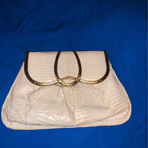 May include: White clutch with a snakeskin pattern and gold-colored accents. The clutch has a flap closure and a gold-colored clasp. The bag is on a blue background.